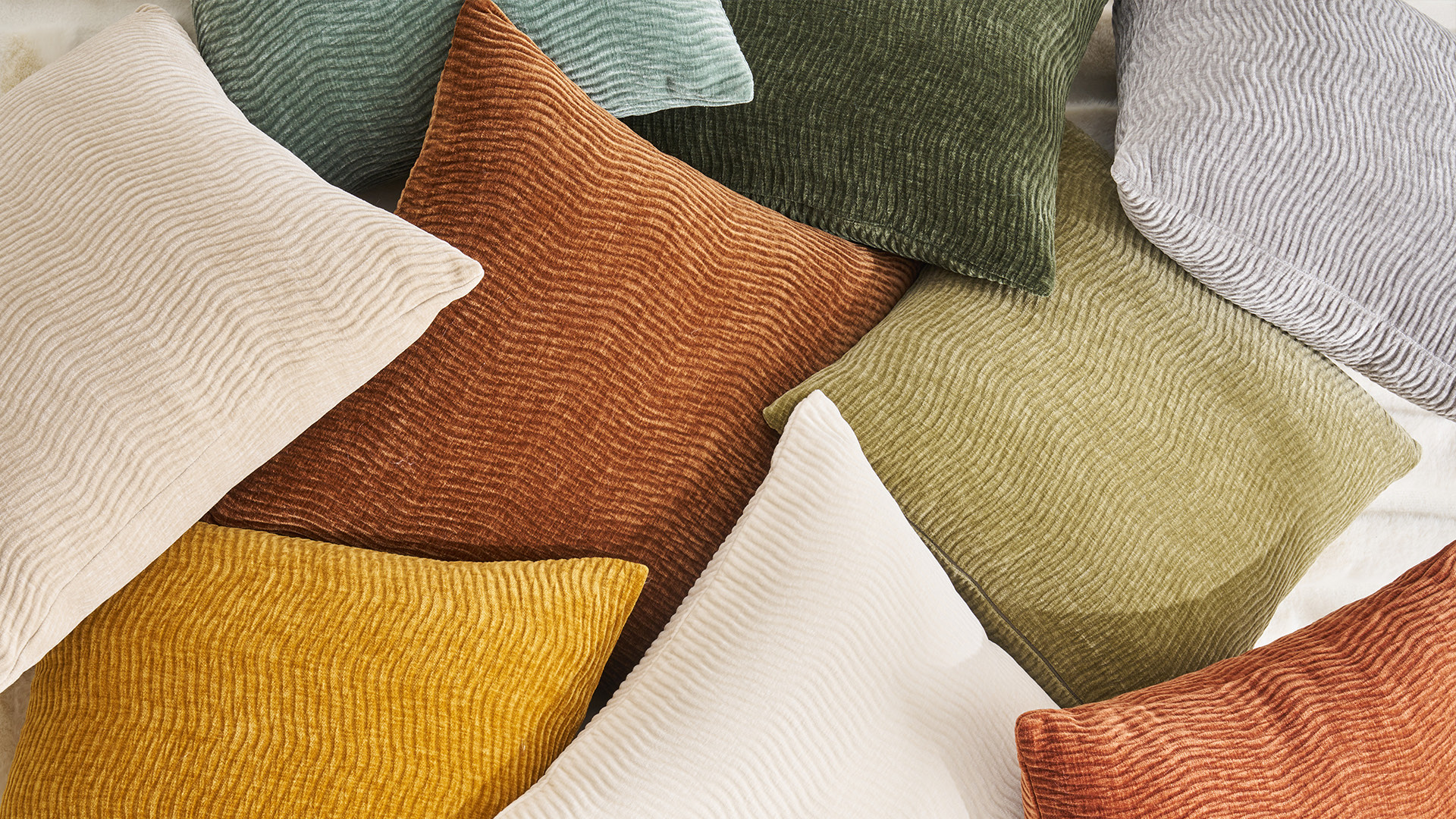 ABS International home textiles — a curated arrangement of textured deco pillows in warm cream, ochre, terracotta and forest-green tones